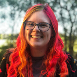 A photograph of a fair skinned person with short brown hair and glasses smiles directly into the camera. They wear a round black hat, a dark green shirt with white flowers, and a blue jean jacket wiht a pin of the right of a shark in space.